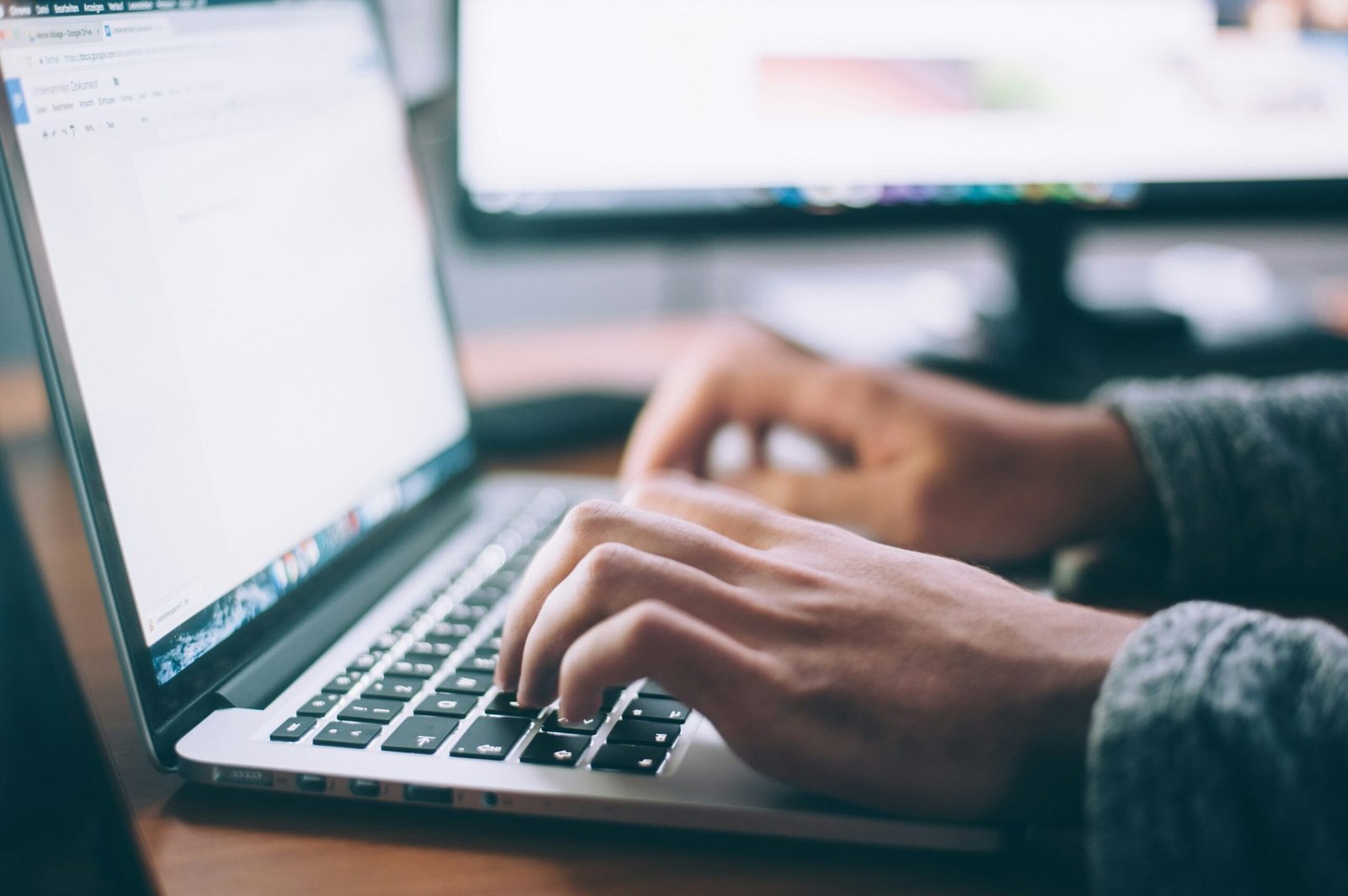 Mãos digitando em um notebook, com um monitor desfocado ao fundo